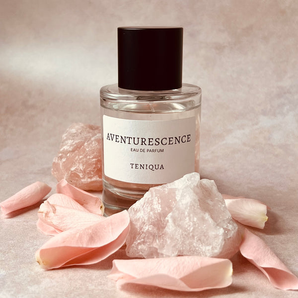 Perfume bottle labeled 'Aventurescence' by Teniqua with pink petals and crystals on a neutral background